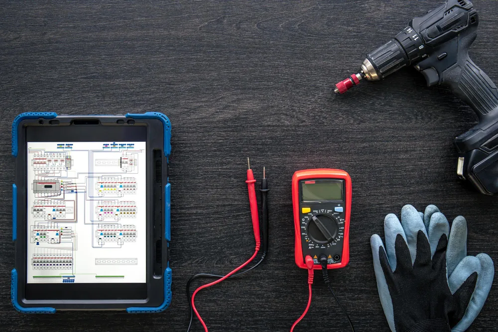 Electrical testing tools, a tablet, work gloves and a drill laid out on a workbench